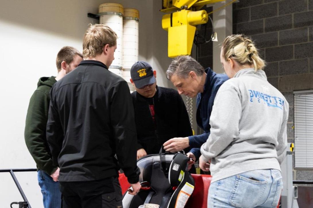 A group of students watch a professor work with a car seat in the Crash Lab